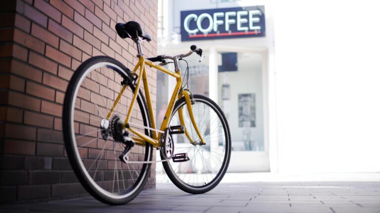 A yellow bicycle is parked on a sidewalk next to a brick wall in front of a white building with a large COFFEE sign above the entrance.