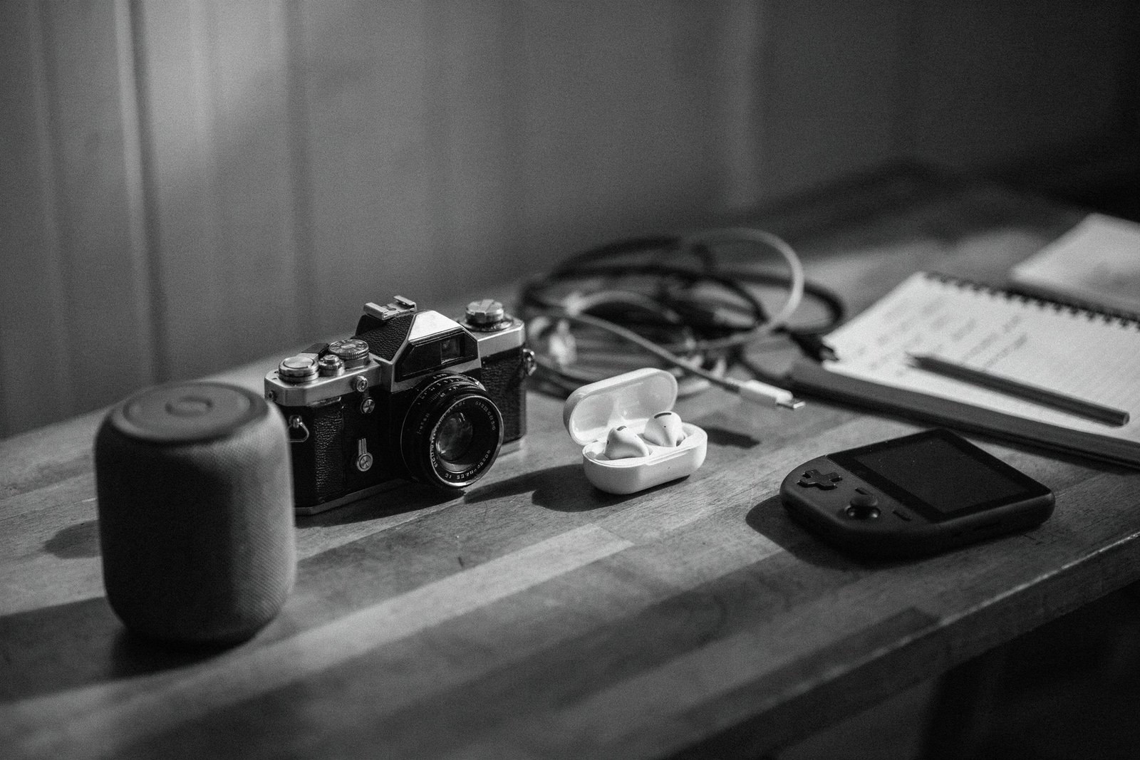 Black and white photo of a reviewer workbench with a film camera, earbuds case, handheld console and notebook